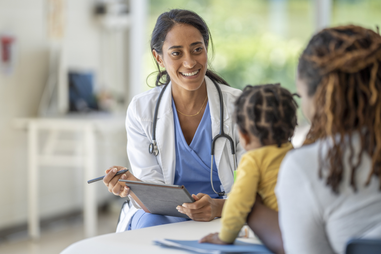 A smiling nurse practitioner talks with a parent and child in an exam room.