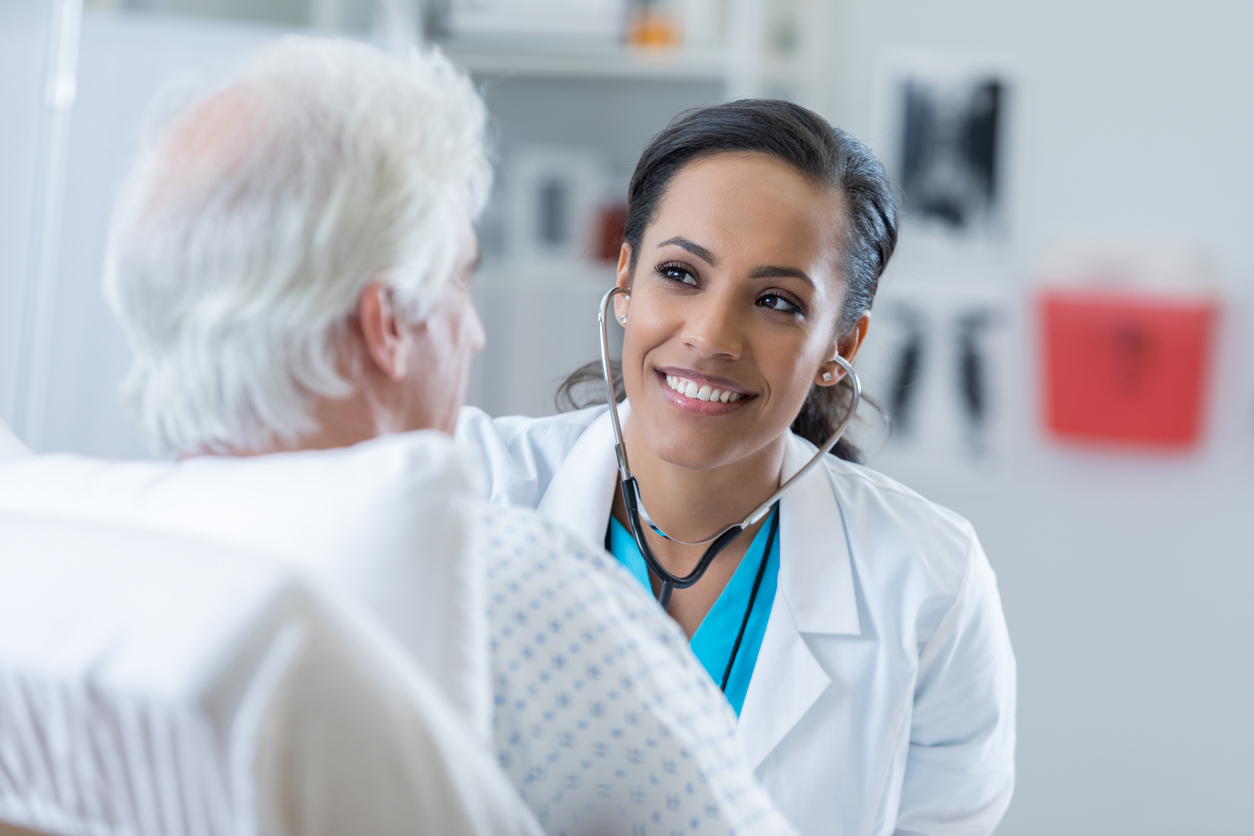 A nurse with a stethoscope listens to the breathing of an older patient in a hospital bed.