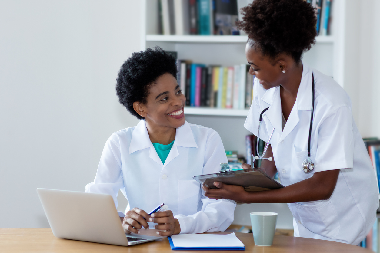 Research nurses in an office discuss a research study while working over a laptop and writing on a clipboard.