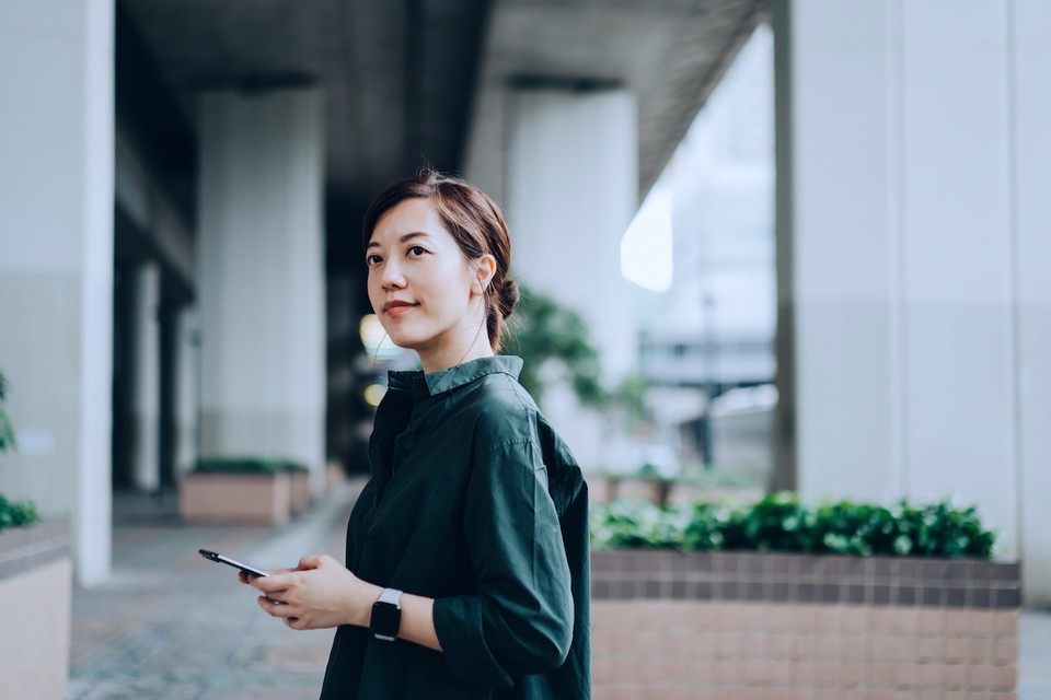 A twenty-something young professional stands and smiles outside of a large office building, wearing a green button-down shirt and holding a mobile phone.