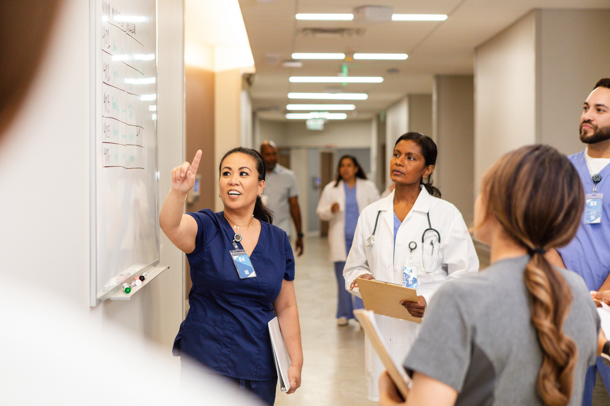 A nurse leader refers to a whiteboard while talking to her team.
